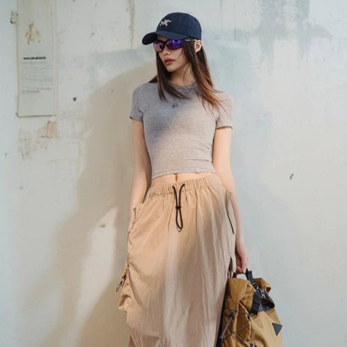 Person wearing a gray knit top, beige pants, and a black cap with sunglasses, standing next to a backpack against a white wall.